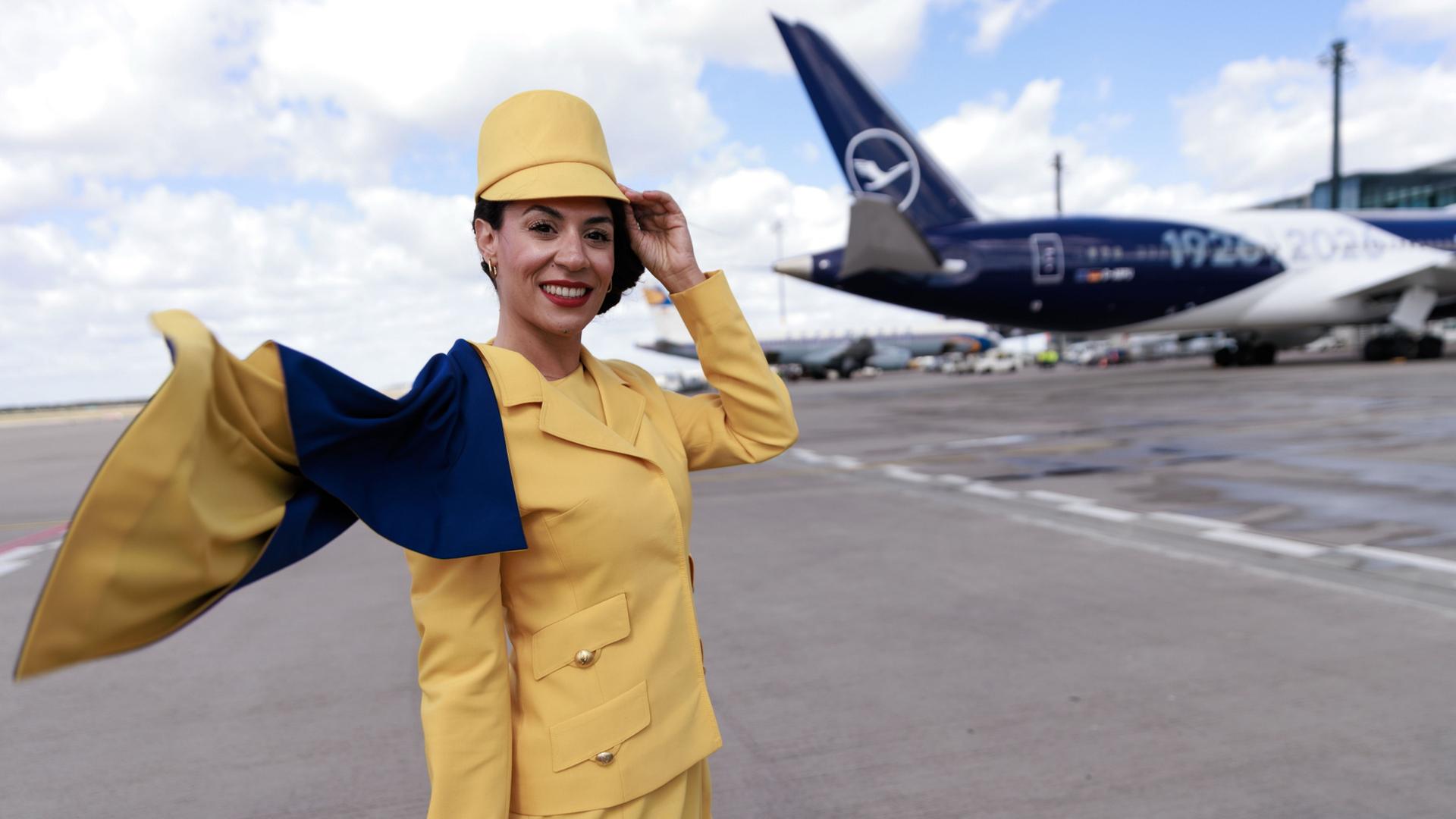 Eine Hostess trägt eine frühere Lufthansa-Uniform vor einem Passagierflugzeug der Lufthansa auf dem Flughafen Berlin-Brandenburg (BER) bei der Feier zum 100. Jahrestag des Erstflugs. | Carsten Koall/dpa