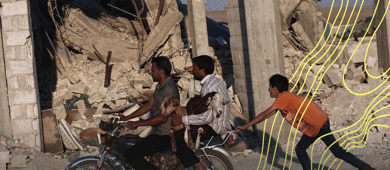 Syrer tragen zwei Ziegen auf einem Motorrad vor zerstörten Gebäuden südlich von Kobane, Syrien.