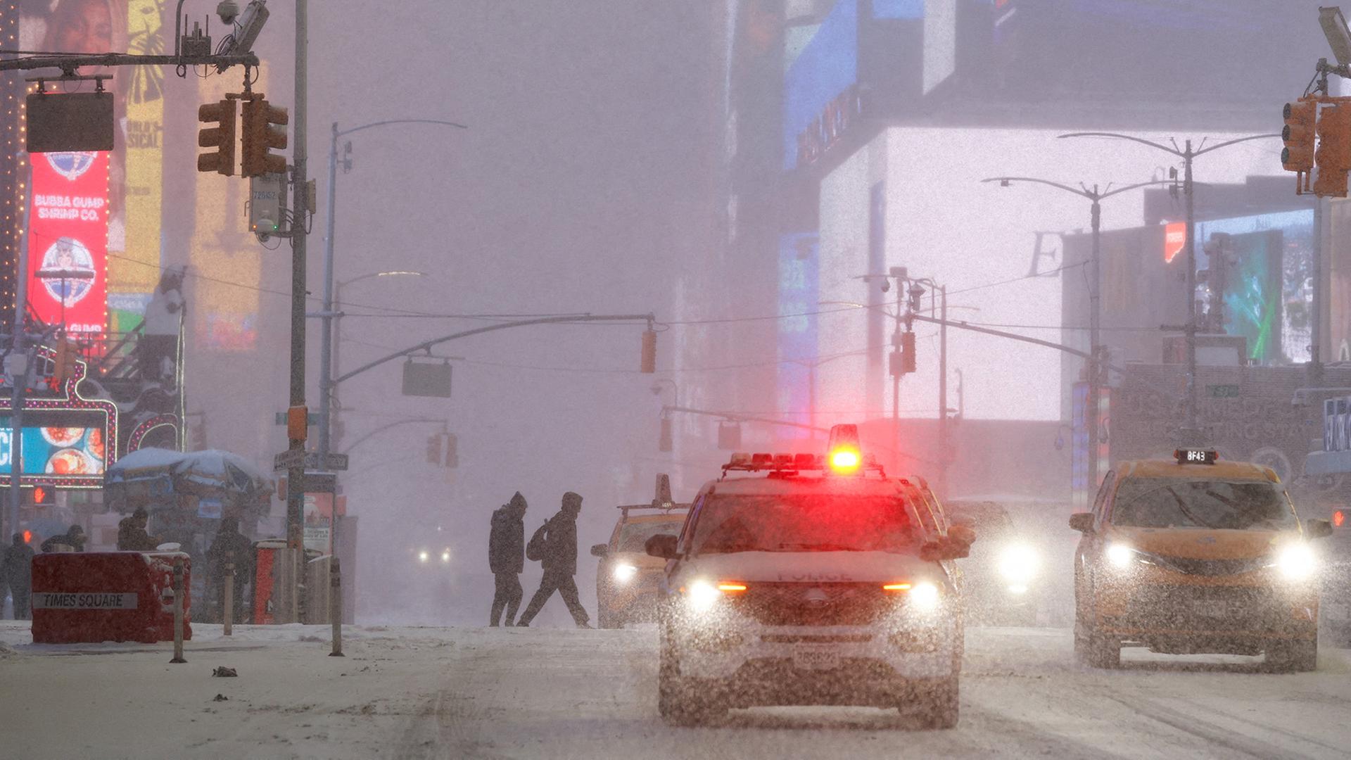 Times Square, New York: Mehrere Menschen überqueren eine verschneite Straße bei starkem Schneefall, ein Polizeiwagen mit eingeschaltetem Blaulicht und ein Taxi fahren durch die Szene.  | REUTERS
