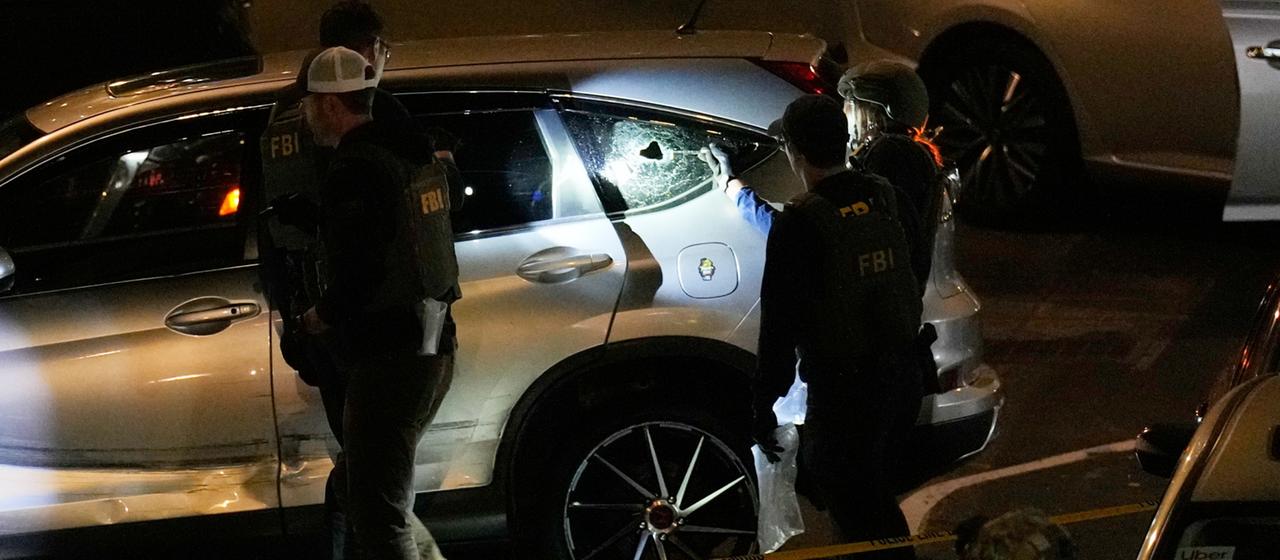 FBI-Agenten untersuchen ein Auto, in der hinteren Scheibe ist ein Einschussloch.