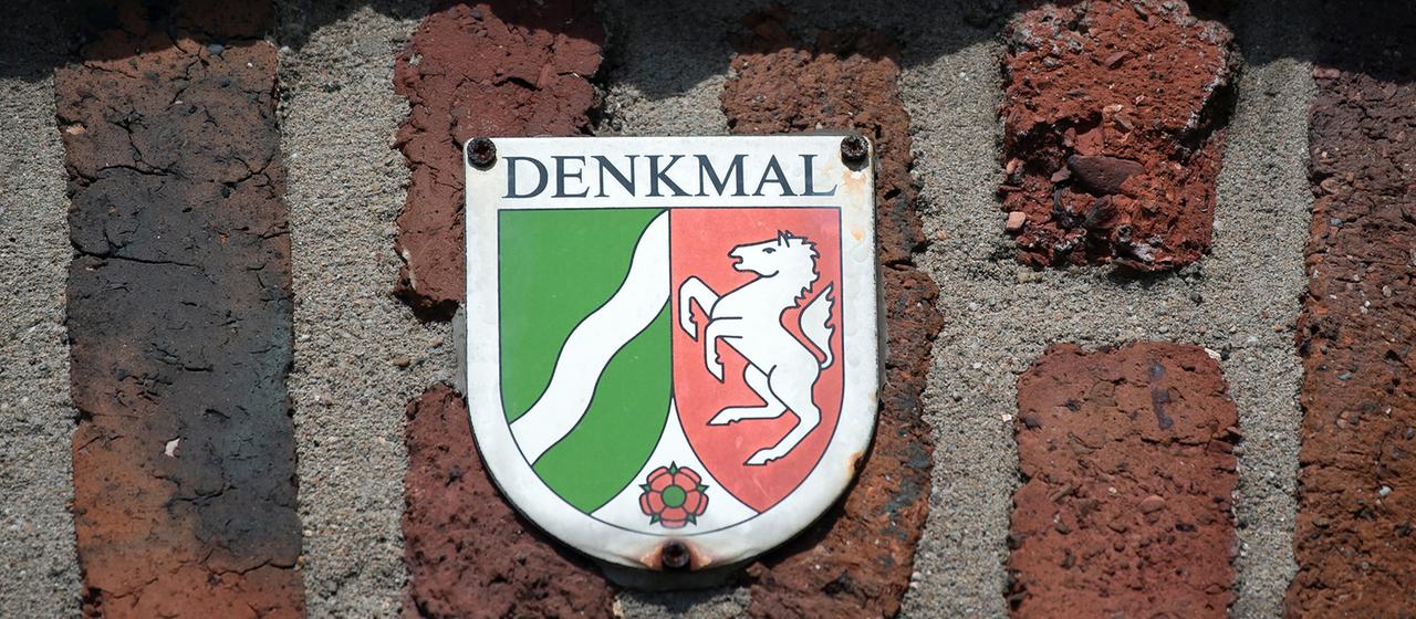 Ein Schild mit der Aufschrift "Denkmal" und dem Landeswappen von Nordrhein-Westfalen hängt an der Wand einer historischen Wohnanlage.