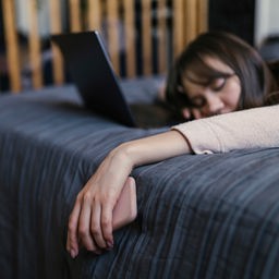 Eine Frau mit einem Handy in der Hand liegt mit geschlossenen Augen vor einem Laptop auf einem Bett.