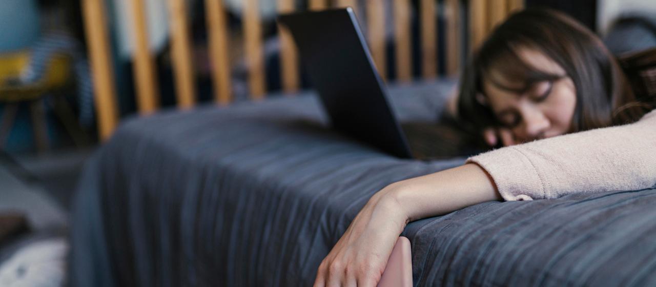 Eine Frau mit einem Handy in der Hand liegt mit geschlossenen Augen vor einem Laptop auf einem Bett.