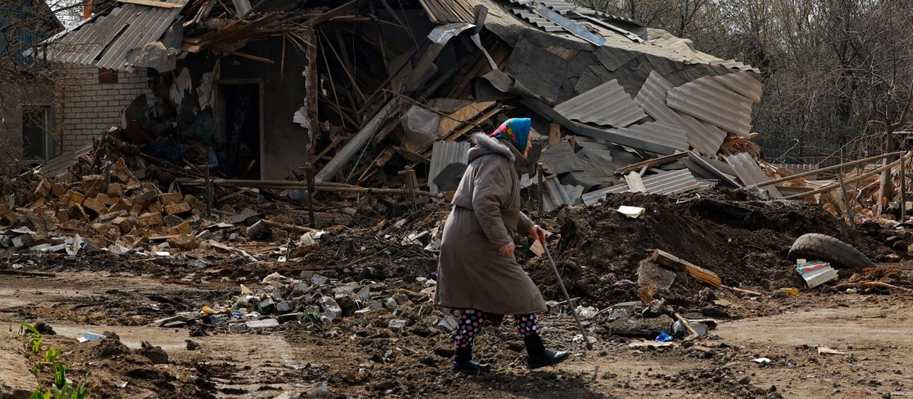 Eine ältere Frau geht vor einem zerstörten Gebäude entlang.