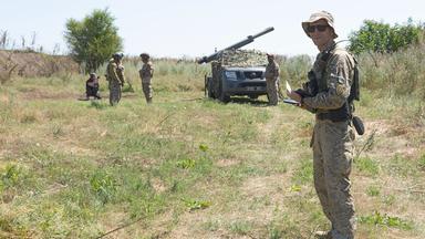 Volodymyr steht auf einer Wiese, im Hintergrund sind Soldaten und ein Raketenwerfer.