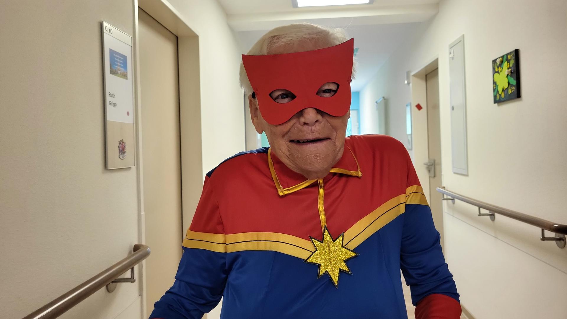 Eine ältere Frau steht in einem Flur und trägt ein rot-blaues Superheldenkostüm mit roter Augenmaske und goldenem Stern. | WDR / Luca Peters