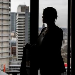 Silhouette einer Frau, die in einem Büro im oberen Stock eines Hochauses aus dem Fenster schaut.