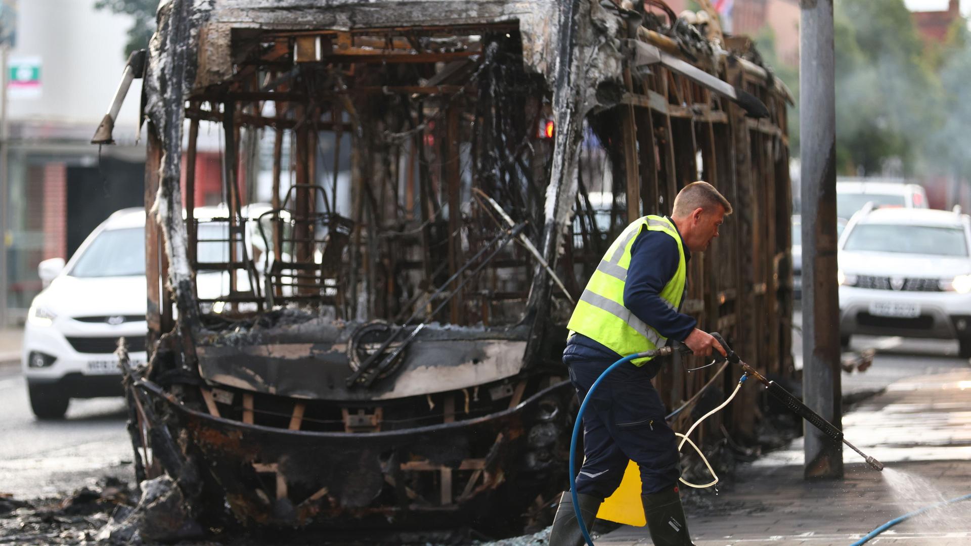 Ein Arbeiter räumt die Trümmer vor einem ausgebrannten Bus auf. | Peter Morrison/AP/dpa
