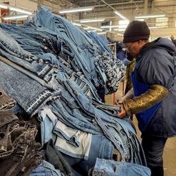 Eine Frau steht in einer Fabrik vor einem Berg aufgestapelter Jeans.