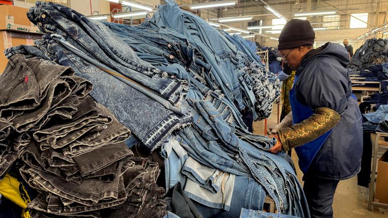 Eine Frau steht in einer Fabrik vor einem Berg aufgestapelter Jeans.