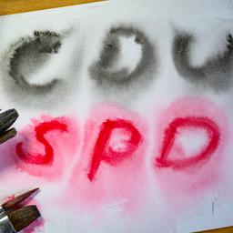 Auf einem Blatt Papier steht CDU in schwarzer und SPD in roter Schrift, daneben liegt ein Malkasten.