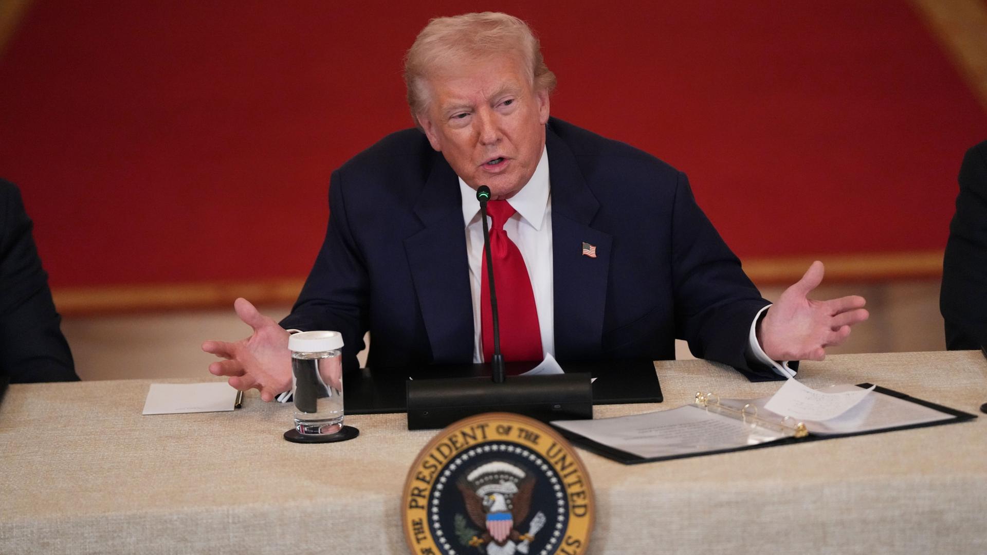 Donald Trump sitzt an einem Tisch vor rotem Hintergrund. | AP