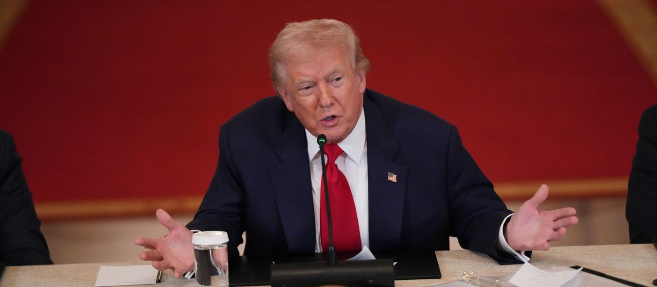 Donald Trump sitzt an einem Tisch vor rotem Hintergrund.