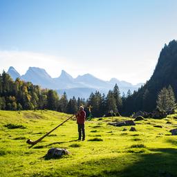 Ein Mann spielt sein Alphorn in freier Natur auf der Alp Laui (Schweiz), im Hintergrund die Bergkette der Churfirsten.