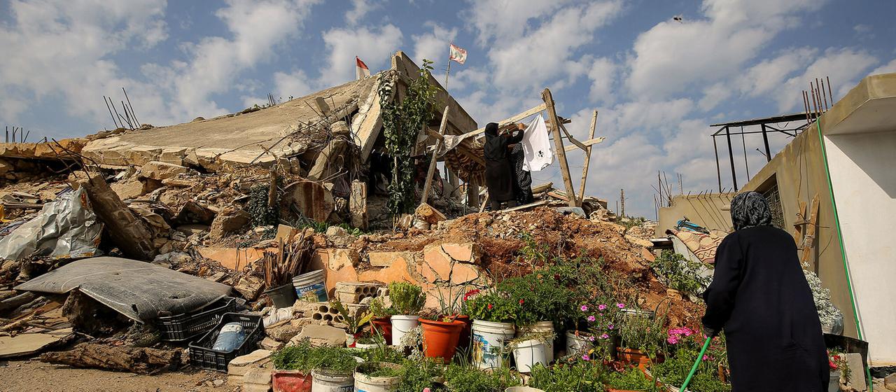 Eine Frau nimmt Wäsche ab, die auf einem Trümmerhaufen aufgehängt ist während eine andere Blumen gießt.