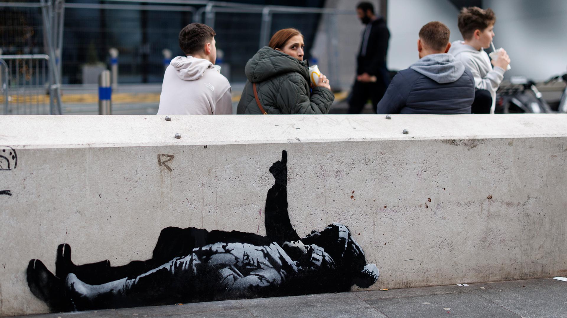 Menschen sitzen hinter einer Mauer, auf der ein Streetartkunstwerk mit zwei liegenden Kindern zu sehen ist. | EPA
