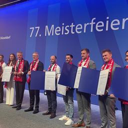 Die Absolventen stehen auf der Bühne in einer Reihe und halten ihre Meisterurkunde in die Kamera