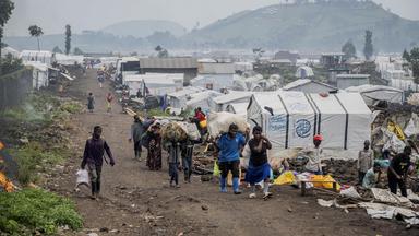 Mehrere Menschen, darunter Kinder, tragen Gepäck durch ein Flüchtlingslager. Im Hintergrund zahlreiche Zelte und provisorische Unterkünfte.
