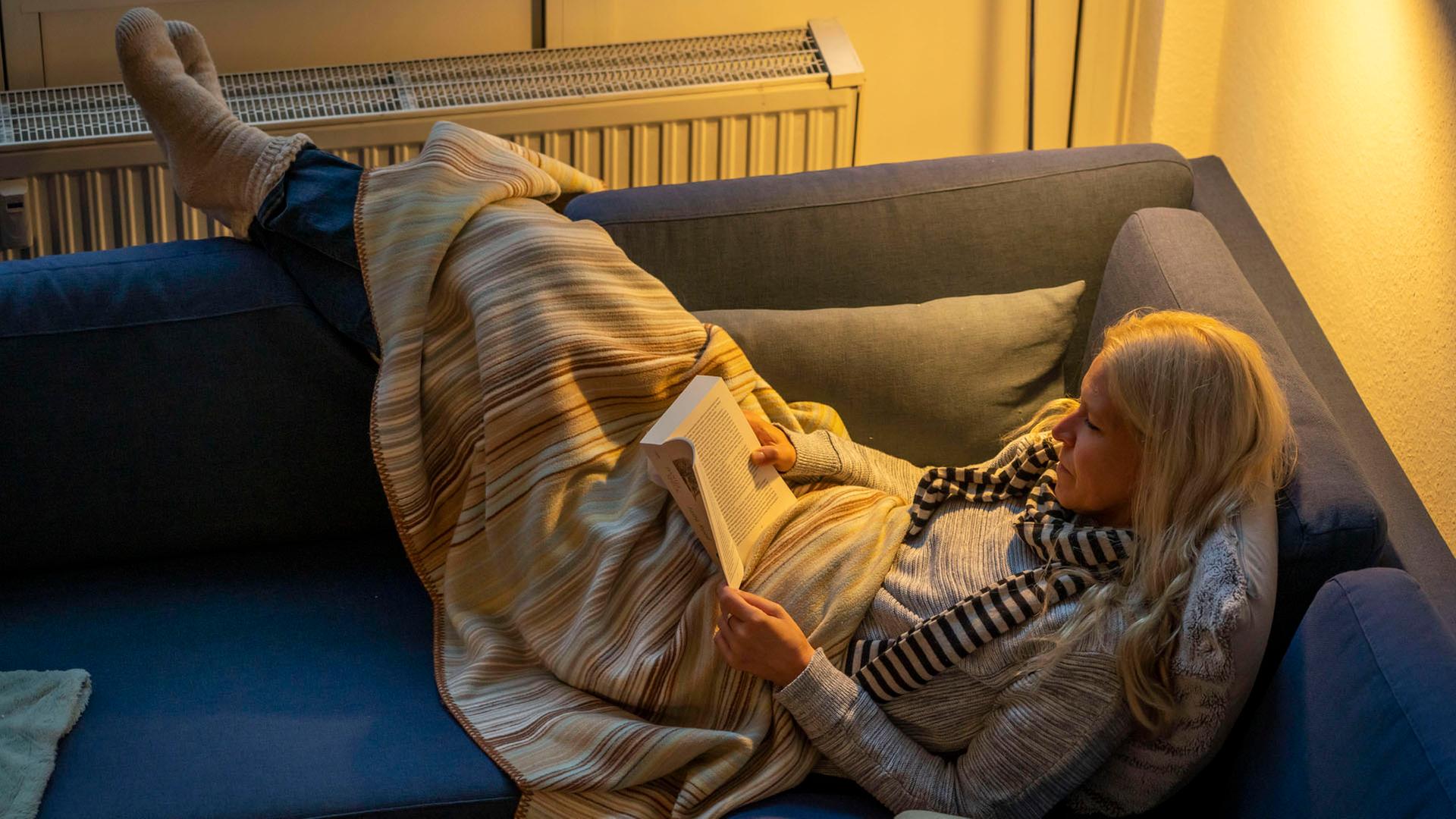 Eine Frau liegt mit einer Wolldecke auf dem Sofa und liest ein Buch. | picture alliance / Jochen Tack