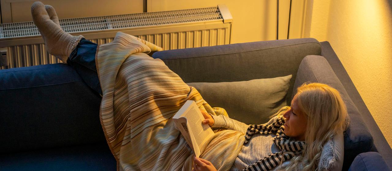 Eine Frau liegt mit einer Wolldecke auf dem Sofa und liest ein Buch.