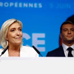 Marine Le Pen blickt lächelnd zur Seite, hinter ihr steht Jordan Bardella.