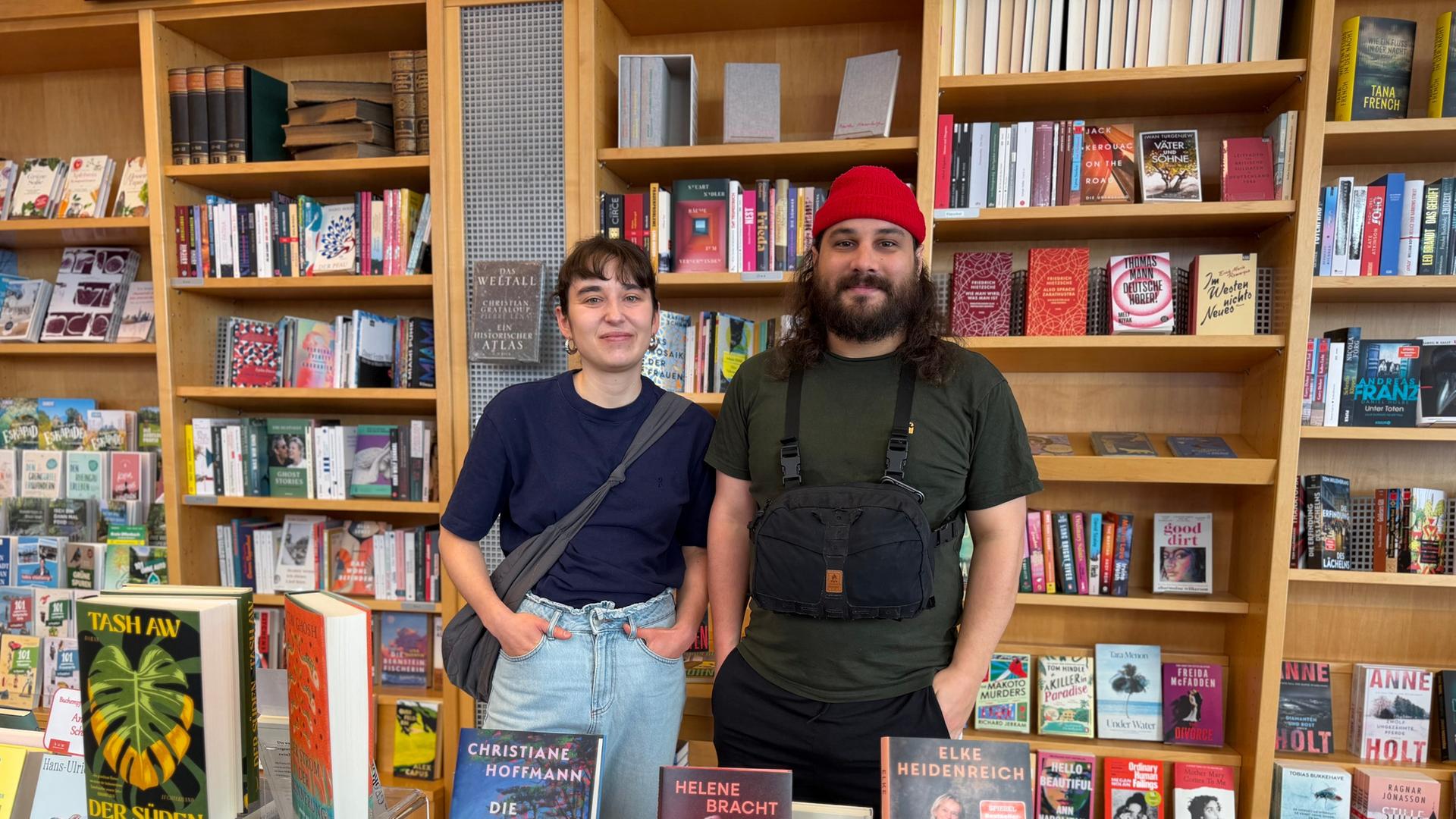 Eine Frau mit blauem T-Shirt und Jeans und ein Mann mit knallroter Mütze und Bart stehen nebeneinander hinter einem Büchertisch und vor einem Bücherregal. | hr/Milena Pieper