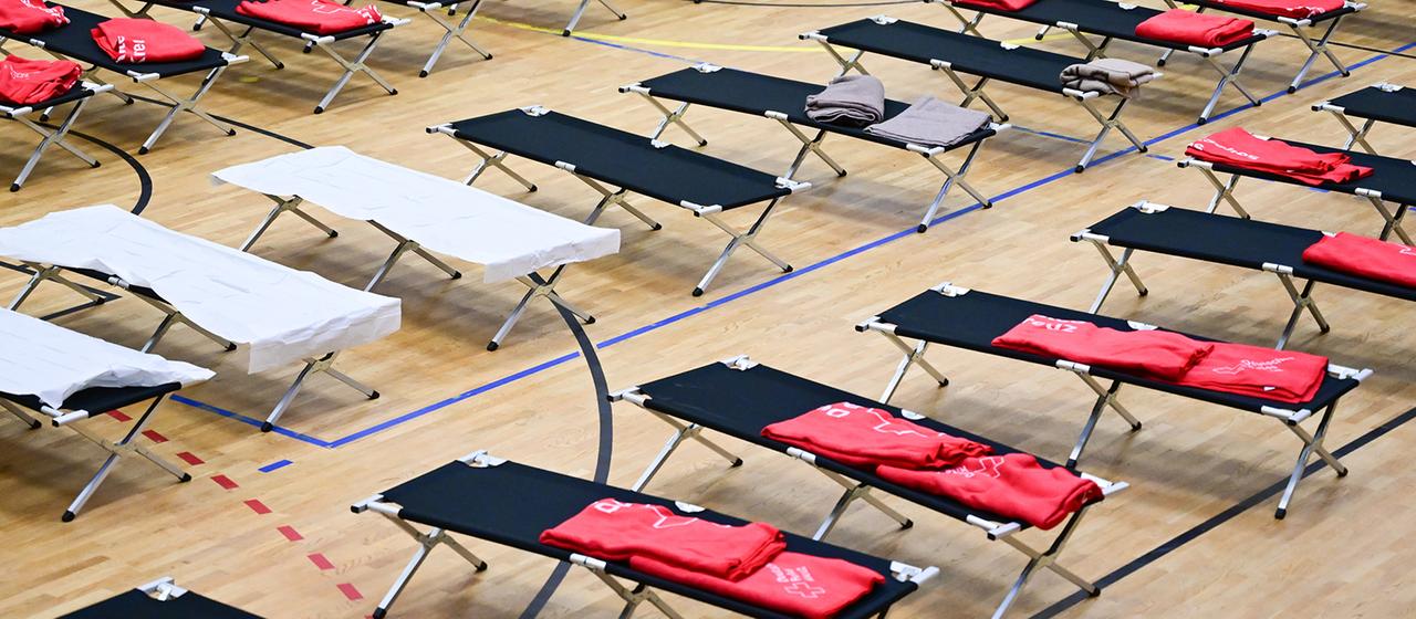 In einer Sporthalle sind zahlreiche Feldbetten aufgestellt.