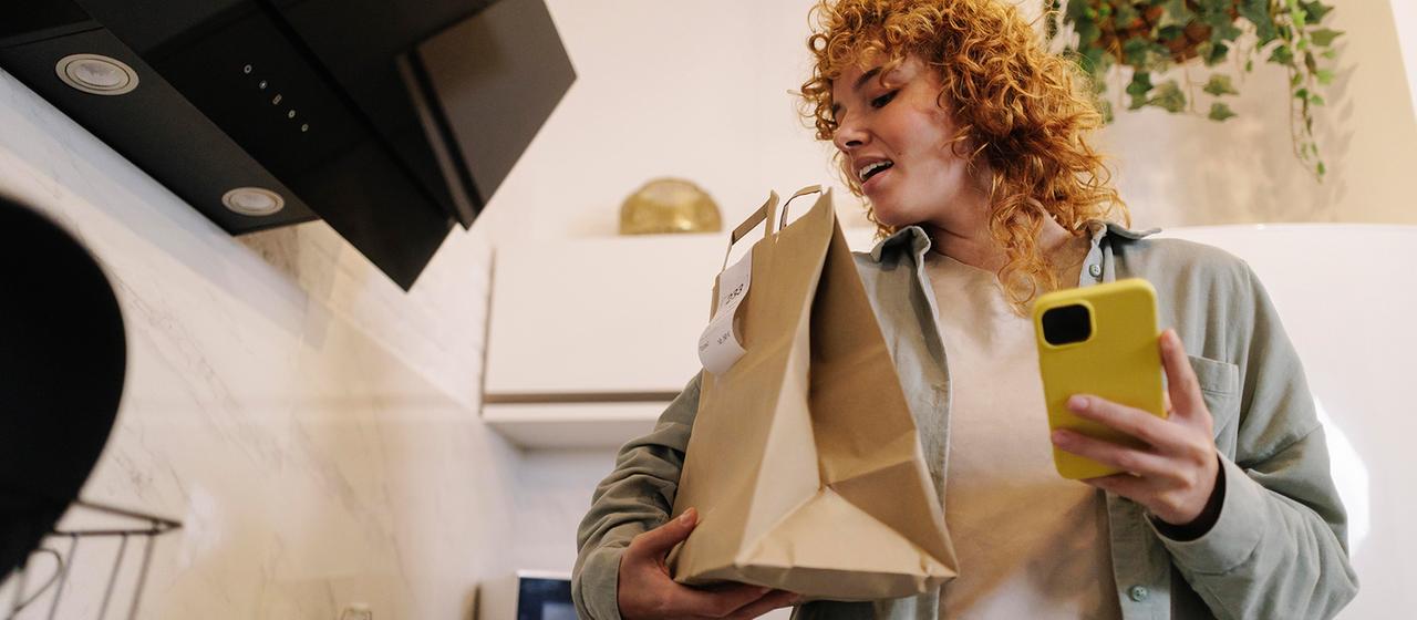 Eine Frau steht mit einer Tüte und einem Handy in der Hand in einer Küche.