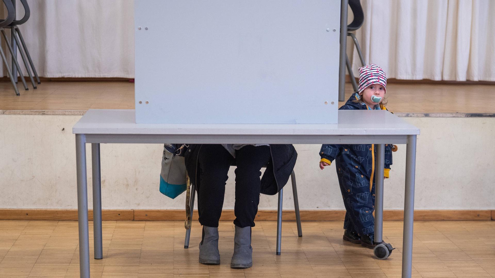 Eine Frau sitzt in einer Wahlkabine. Man sieht nur ihre Beine, wÃ¤hrend sie hinter einer Sichtblende sitzt. Neben dem Tisch steht ein kleines Kind und schaut neugierig zur Kamera. | IMAGO / EHL Media