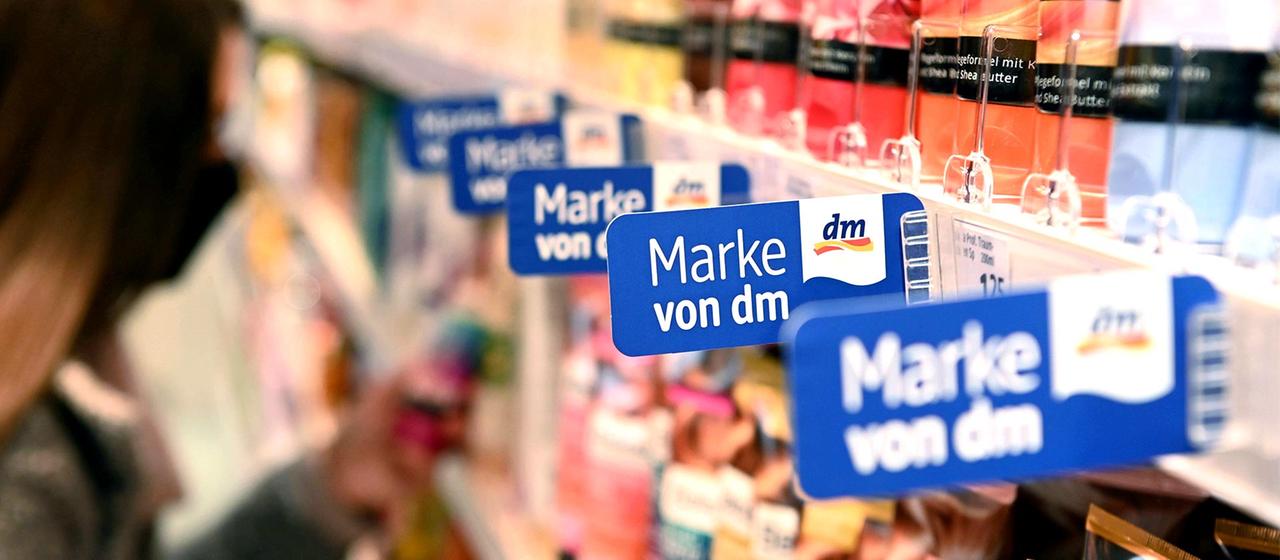 In einem Regal einer Filiale der Drogeriemarktkette dm hängen Schilder, die auf die Eigenmarken hin weisen.