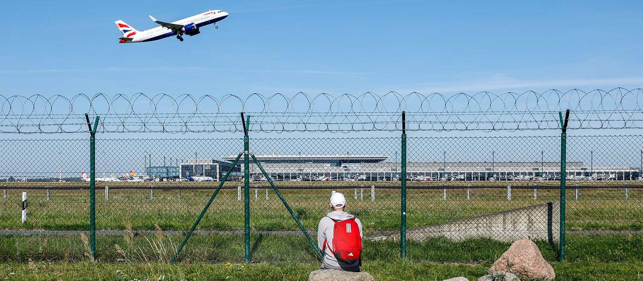 Ein Mann beobachtet durch einen Zaun den Start eines Flugzeugs am Flughafen BER.