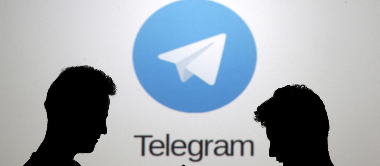 Zwei Männer posieren mit Smartphones vor einem Bildschirm, auf dem das Telegram-Logo zu sehen ist.