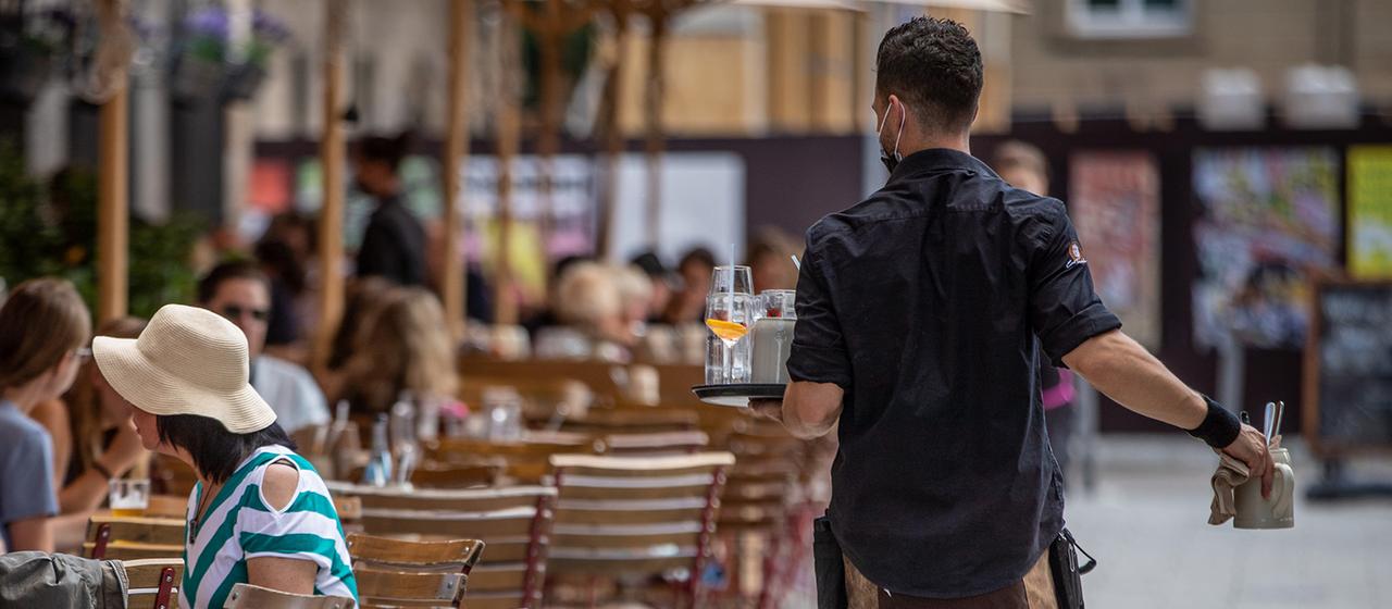 Eine Servicekraft läuft im Außenbereich eines Restaurants an Gästen vorbei.