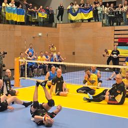 Zwei Mannschaften mit jeweils 6 Personen sitzen durch ein tiefes Volleyball- Netz getrennt. Näher an der Kamera sitzt das schwarz- tragende deutsche Team, weiter hinten die Gelb- tragenden Ukrainer. 