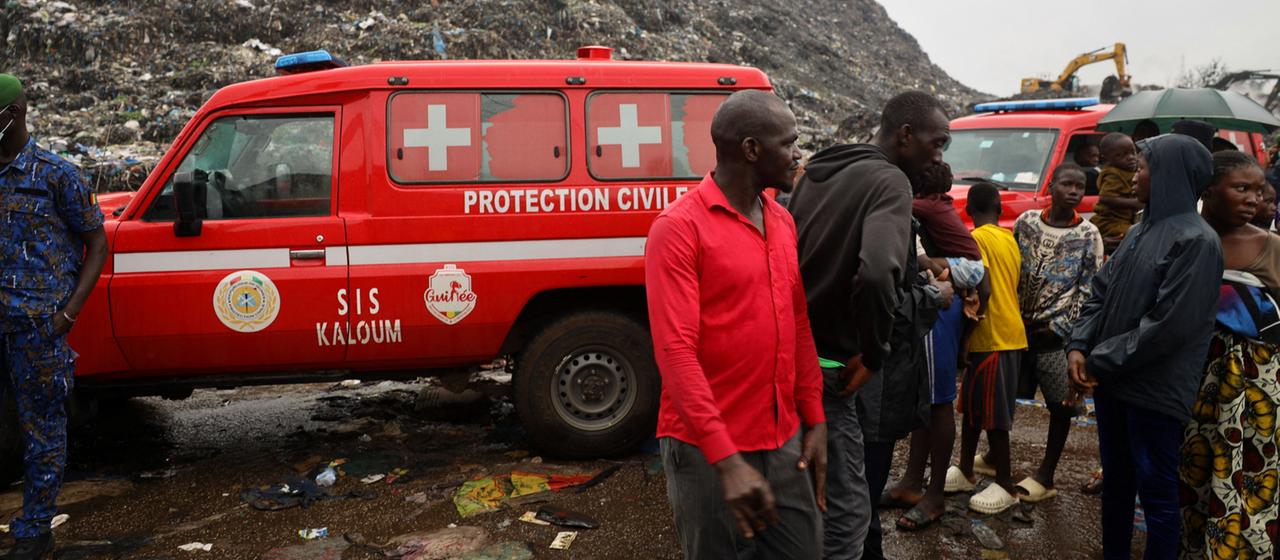 Menschen stehen vor roten Rettungswagen auf einer Mülldeponie in Guinea