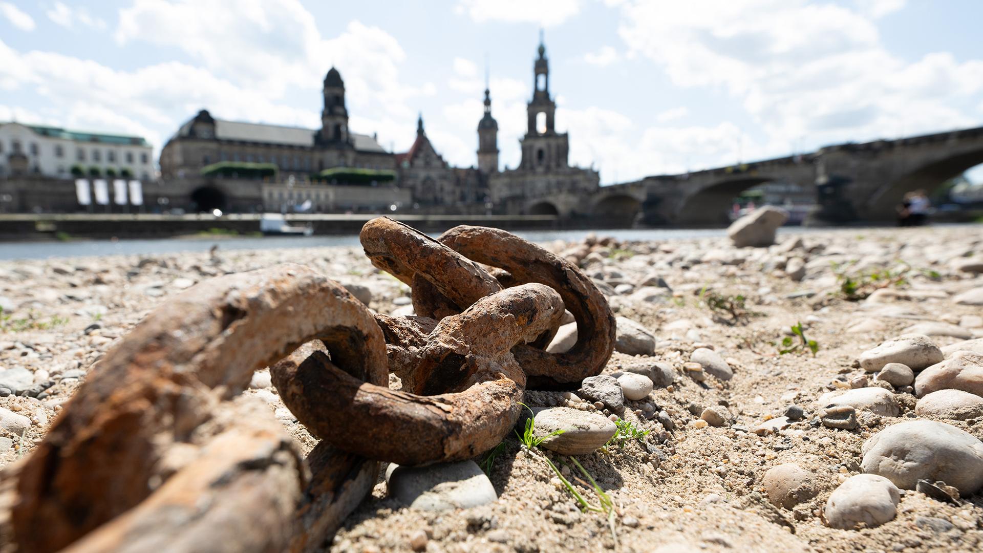  Eine rostige Kette liegt im ausgetrockneten Flussbett am Ufer einer Stadt. (Archivbild: 26.6.2025) | dpa