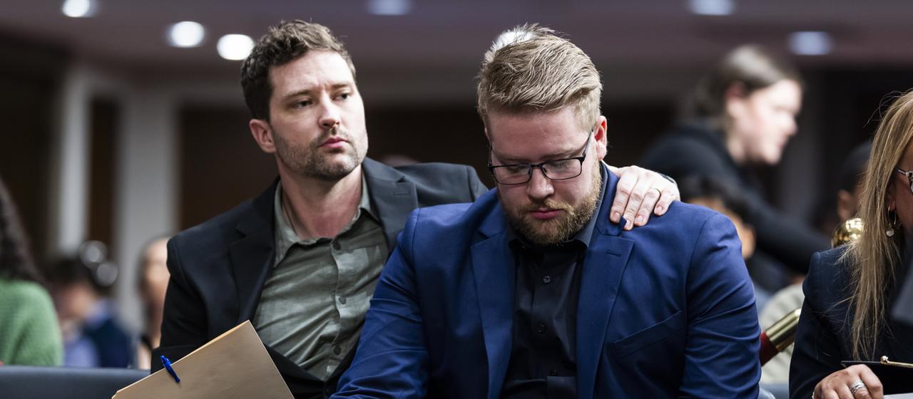 Brent und Luke Ganger sitzen auf Stühlen, Luke hält den Kopf gesenkt, sein Bruder hat einen Arm um seine Schultern gelegt.