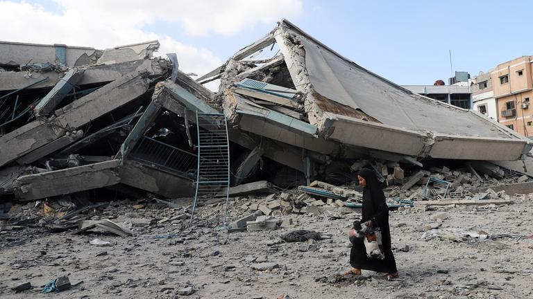 Eine Frau trägt Gegenstände vor einem zerstörten Gebäude.