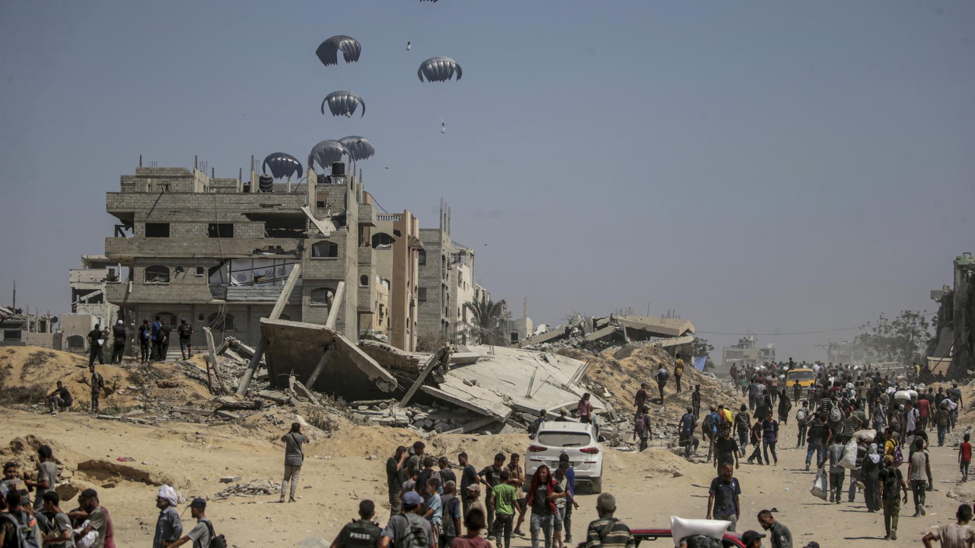 Humanitäre Hilfe landet mit Fallschirmen im Gazastreifen, davor stehen Menschen. | EPA
