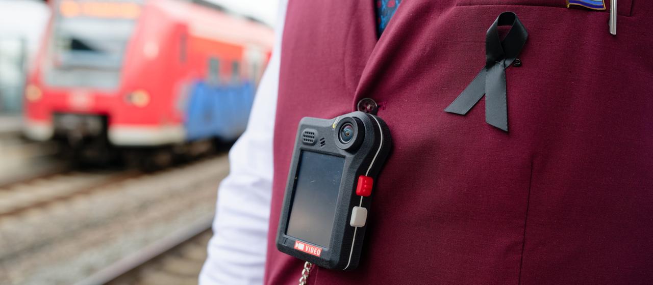 Ein Zugbegleiter mit einer Bodycam und schwarzer Schleife vor einem roten Zug