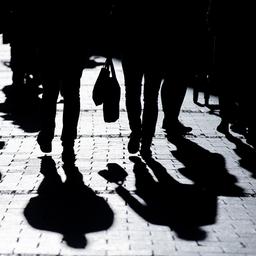 Passanten gehen in starkem Gegenlicht durch eine Fußgängerzone und werfen Schatten auf den Asphalt. 