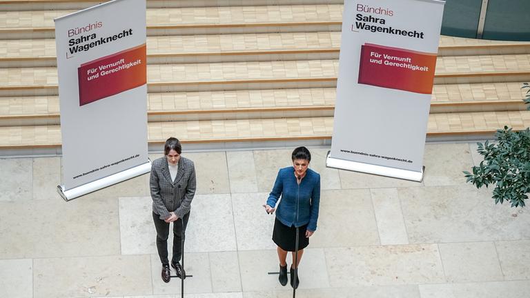 Plakate des Bündnisses Sahra Wagenknecht hängen von der Decke, darunter stehen Sahra Wagenknecht und ihre Sprecherin 