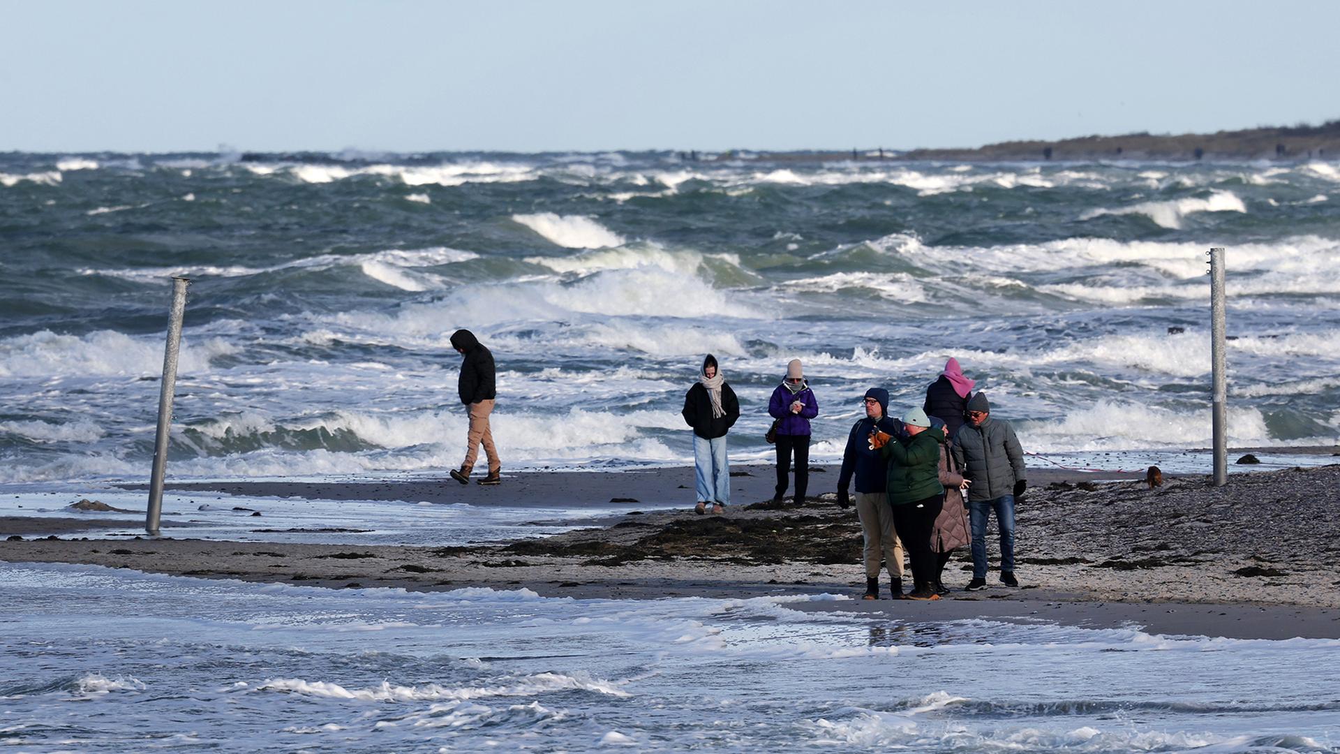 Menschen spazieren bei starker Brandung an einem Strand. | dpa