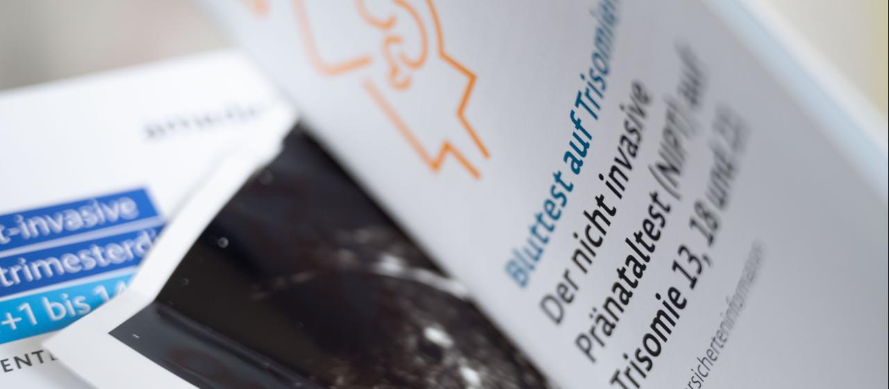 Zwei Informationsbroschüren zu nichtinvasiven Pränataltests liegen neben einem Ultraschallbild in einer Frauenarztpraxis.