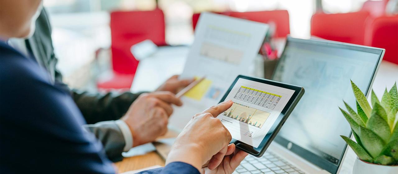 Zwei Personen betrachten Finanzdaten auf Tablet, Laptop und Papier.