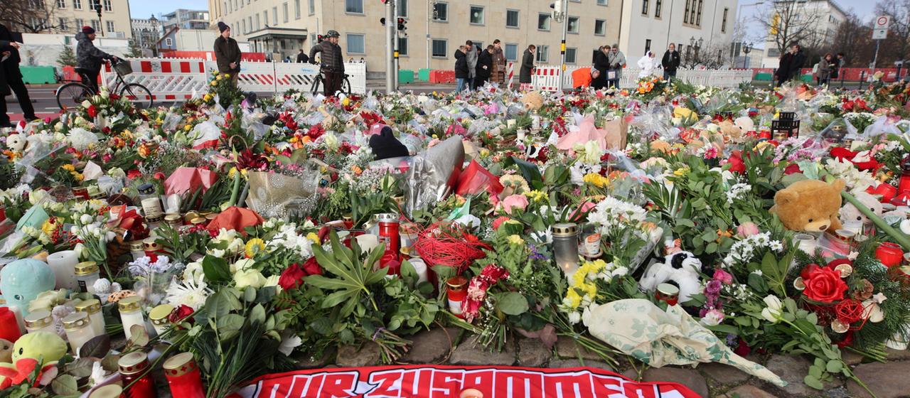 Menschen stehen am Gedenkort zum Anschlag in Magdeburg vor einem Meer aus Blumen, Kerzen und Plüschtieren