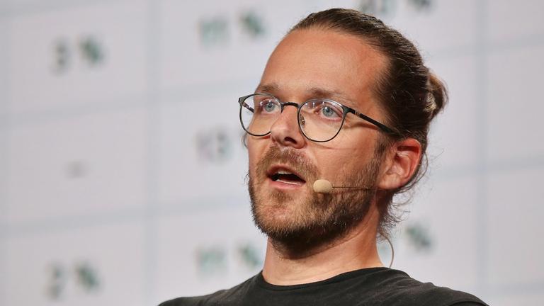 Arne Semsrott: Junger Mann mit kurzem Vollbart, braunen Haaren zum Zopf zurückgebunden, Brille und blauen Augen.