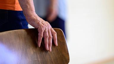 Eine alte Frau stützt sich auf einen Stuhl.