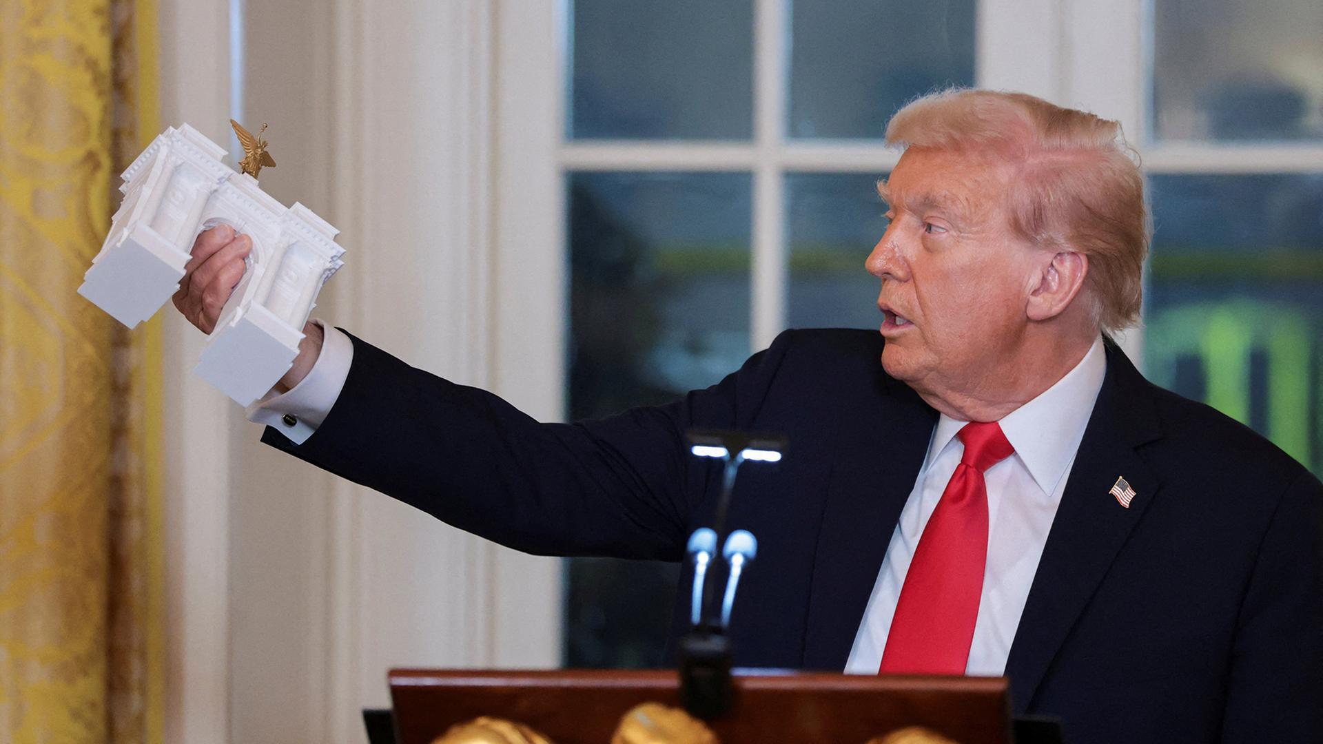 Donald Trump hält ein Modell eines monumentalen Bogens. | REUTERS