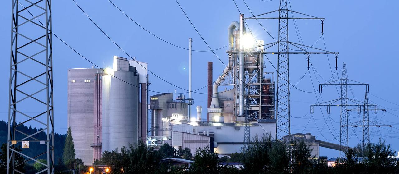 Strommasten stehen im Morgenlicht vor einem Zementwerk.